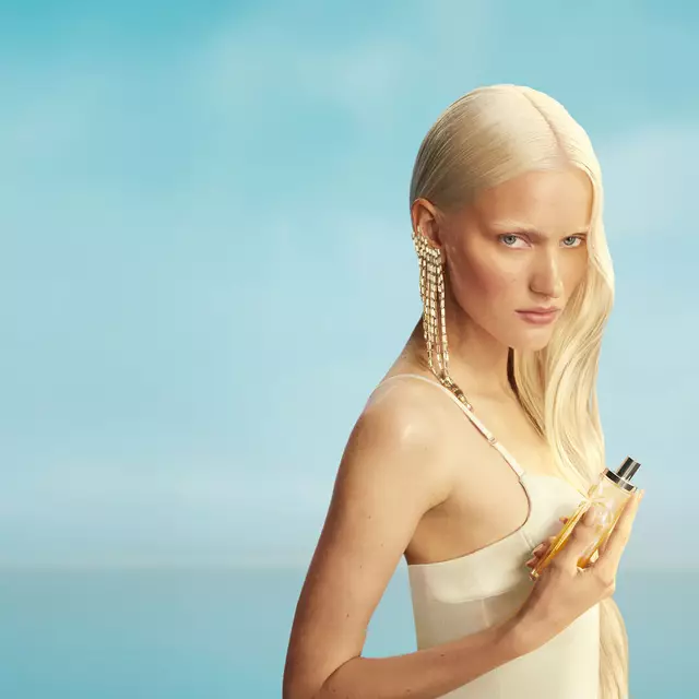 Nuovo banner della campagna Mugler Starlicious les eaux gourmandes con una modella con lunghi capelli biondi e occhi azzurri, che indossa un vestito giallo pallido, con in mano il flacone della fragranza Pineapple Musk su uno sfondo di cielo azzurro tenue. Le fragranze sono deliziose, da collezione e stratificabili.