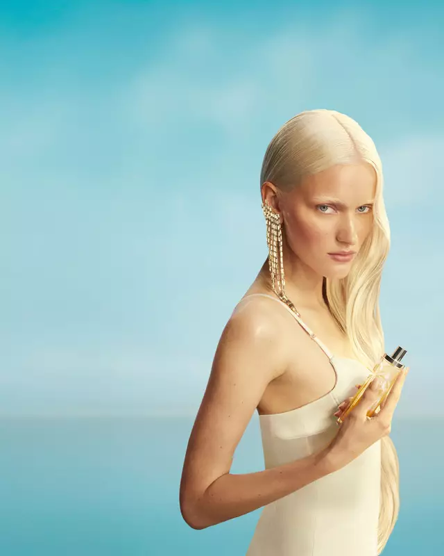 Nuovo banner della campagna Mugler Starlicious les eaux gourmandes con una modella con lunghi capelli biondi e occhi azzurri, che indossa un vestito giallo pallido, con in mano il flacone della fragranza Pineapple Musk su uno sfondo di cielo azzurro tenue. Le fragranze sono deliziose, da collezione e stratificabili.
